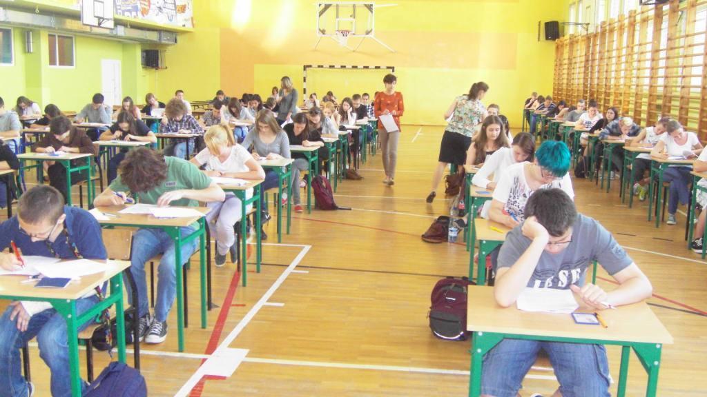 Uczniowie piszą egzamin w sali gimnastycznej w Łomży. Widoczny rząd ławek, uczniowie skupieni na testach, nauczyciele nadzorujący i kosz do koszykówki na ścianie.