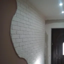 Ściana z dekoracyjnej cegły pomalowanej na biało, z fragmentem ściany wykończonej gładzią w kolorze beżowym, widoczne oświetlenie sufitowe i fragment drzwi.