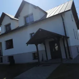 Dwukondygnacyjny dom z jasną elewacją, ciemnym dachem i drewnianą wiatą nad wejściem, widoczny z perspektywy trawnika w słoneczny dzień.