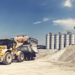 Żółta ładowarka kołowa Hyundai HL760-7A nabiera żwir do naczepy Schmitz, w tle silosy magazynowe i błękitne niebo z chmurami.