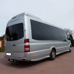 Srebrny, wysoki bus z przyciemnianymi szybami, widok z tyłu i boku, zaparkowany na czerwonej kostce brukowej, z hakiem holowniczym i widocznym logo Mercedesa.