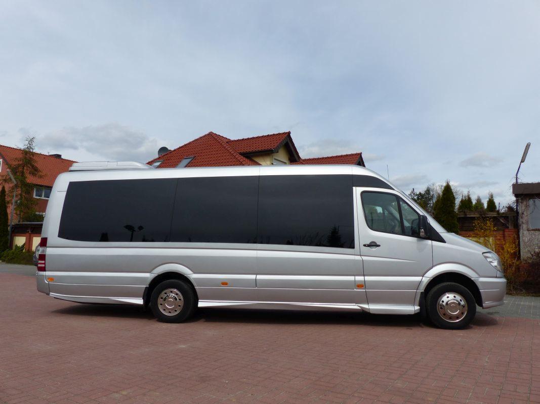 Srebrny, wydłużony bus z przyciemnianymi szybami, zaparkowany na czerwonej kostce brukowej, z budynkiem w tle.