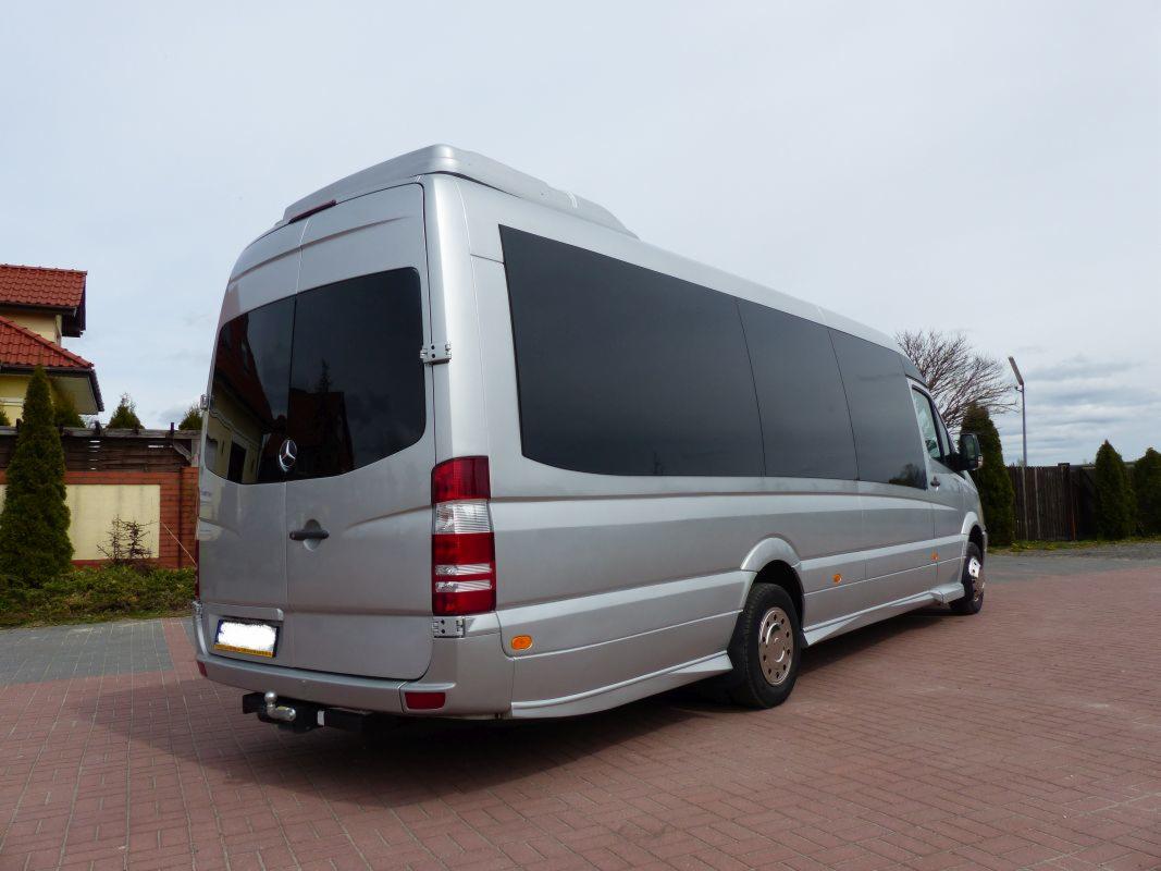 Srebrny, wysoki bus z przyciemnianymi szybami, widok z tyłu i boku, zaparkowany na czerwonej kostce brukowej, z hakiem holowniczym i widocznym logo Mercedesa.