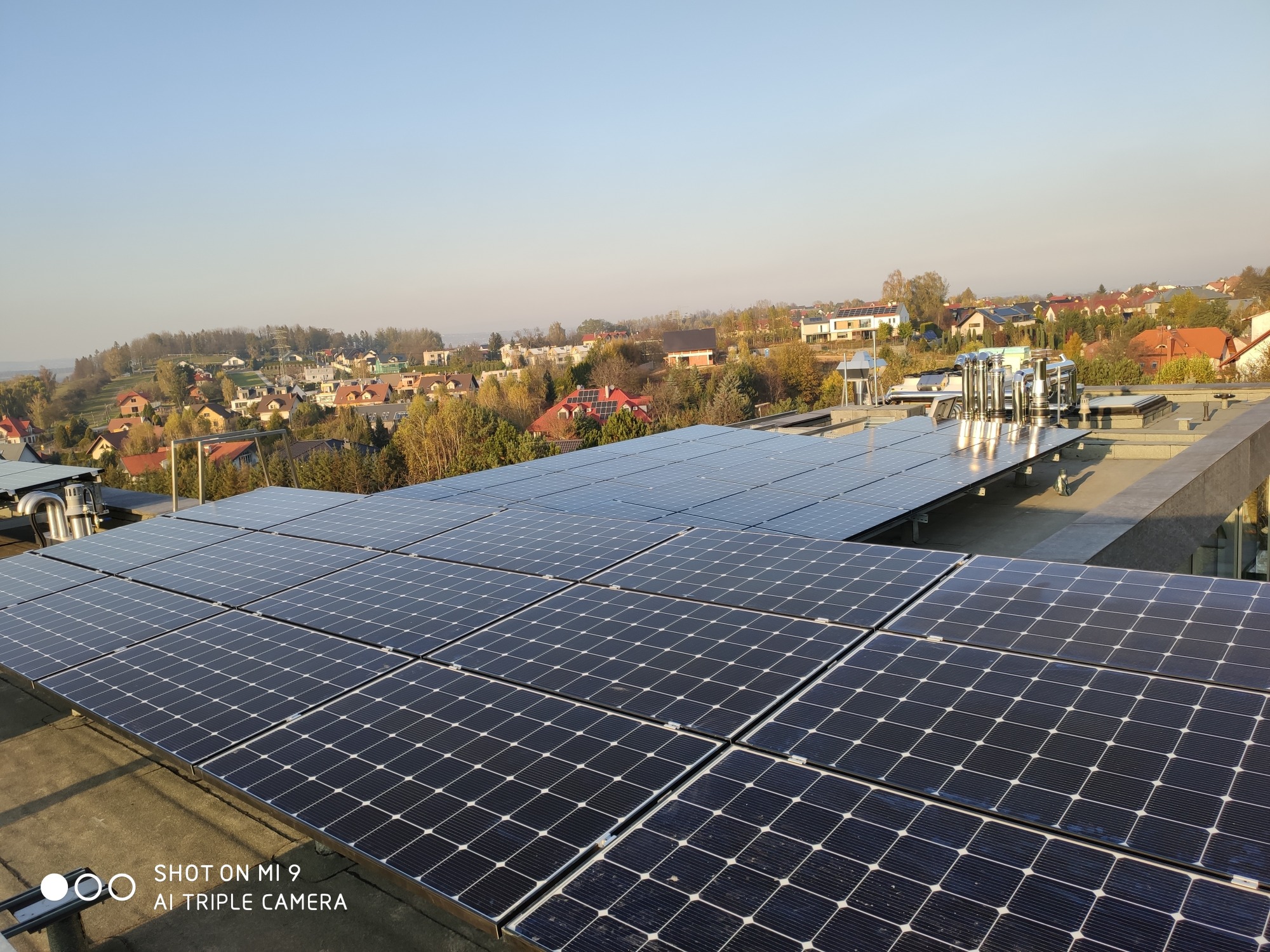 Rozległa instalacja paneli słonecznych na płaskim dachu budynku, z widokiem na zabudowania mieszkalne i drzewa w oddali, sfotografowana w słoneczny dzień.