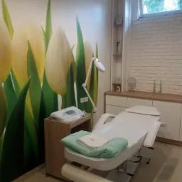 Jasne wnętrze gabinetu kosmetycznego z łóżkiem do zabiegów, aparaturą na drewnianym stoliku, ścianą z fototapetą z motywem tulipanów i dekoracyjną ścianą z białej cegły.