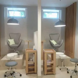 Dwa stanowiska do pedicure z szarymi fotelami, białymi misami do moczenia stóp, drewnianymi regałami i lampami wiszącymi w jasnym wnętrzu z drewnianymi panelami.