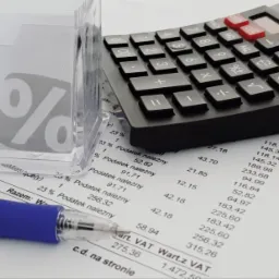 Kalkulator, długopis i dokument z wyliczeniami podatku VAT na biurku, obok wizytówka biura rachunkowego Marzena Rączka w przezroczystym stojaku.