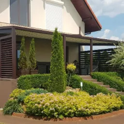 Zadaszony taras z brązowymi lamelami i schodami, otoczony zadbanym ogrodem z różnorodną roślinnością i strzyżonymi krzewami.