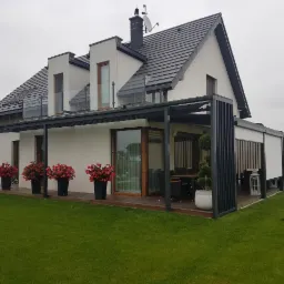 Nowoczesny dom z szarą pergolą z pionowymi panelami, przeszklonym balkonem i tarasem z drewnianą podłogą, ozdobiony czerwonymi kwiatami w czarnych donicach i kulistymi krzewami.