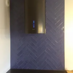 Ciemnoniebieska ściana z płytek ułożonych w jodełkę, nad czarnym blatem, z zamontowanym czarnym okapem z podświetleniem LED.