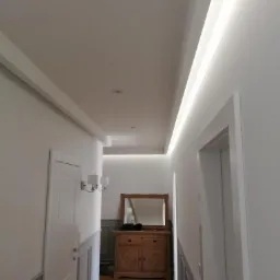 Korytarz z białymi ścianami, sztukaterią, oświetleniem LED w suficie podwieszanym i drewnianą komodą z lustrem w tle, widziany z dołu.