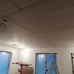 Montaż sufitu podwieszanego z płyt kartonowo-gipsowych, widoczne profile, otwory na oświetlenie i pracownik w tle.