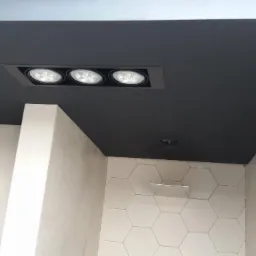 Czarne, matowe wykończenie sufitu z wbudowanym oświetleniem punktowym, kontrastujące z jasnymi ścianami z elementami heksagonalnych płytek.