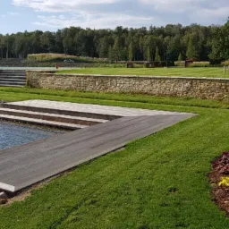 Nowoczesny taras nad jeziorem z drewnianymi schodami prowadzącymi do wody, otoczony kamiennym murem i zadbanym trawnikiem.