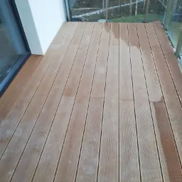 Deski kompozytowe ułożone na balkonie w Olsztynie, widoczne ślady po deszczu i zarys drzew za szklaną balustradą.