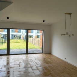 Usługi Ogólnobudowlane Mariusz Filip - Wnętrze jasnego pokoju z dużym oknem tarasowym wychodzącym na ogród, drewnopodobne płytki podłogowe z widocznymi plamami, nowoczesna lampa wisząca z kulistymi kloszami.