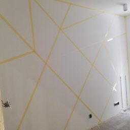 Usługi Ogólnobudowlane Mariusz Filip - Biała ściana z geometrycznym wzorem wyznaczonym taśmą malarską, widoczne puszki elektryczne i ślady po wiązce lasera, przygotowanie do malowania wzoru.