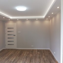 Usługi Ogólnobudowlane Mariusz Filip - Pusty pokój po remoncie z panelami podłogowymi imitującymi drewno, szarą ścianą, białymi drzwiami z przeszkleniami, oświetleniem sufitowym punktowym i centralnym.
