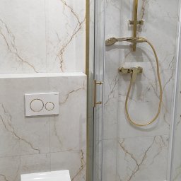 Usługi Ogólnobudowlane Mariusz Filip - Nowoczesna łazienka z marmurowymi płytkami, kabiną prysznicową z złotymi detalami i podwieszaną toaletą z przyciskiem spłukującym w kolorze białym ze złotymi obwódkami.