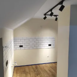 Wykończona łazienka na poddaszu: białe, prostokątne płytki ułożone do połowy ściany, drewnopodobna podłoga z gresu, czarne reflektory sufitowe na szynoprzewodzie.