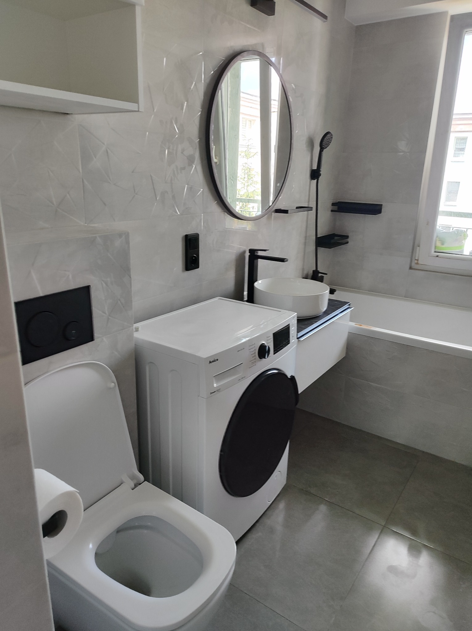 Nowoczesna łazienka z białą armaturą, pralką Amica, okrągłym lustrem i geometrycznymi płytkami ściennymi w odcieniach szarości.