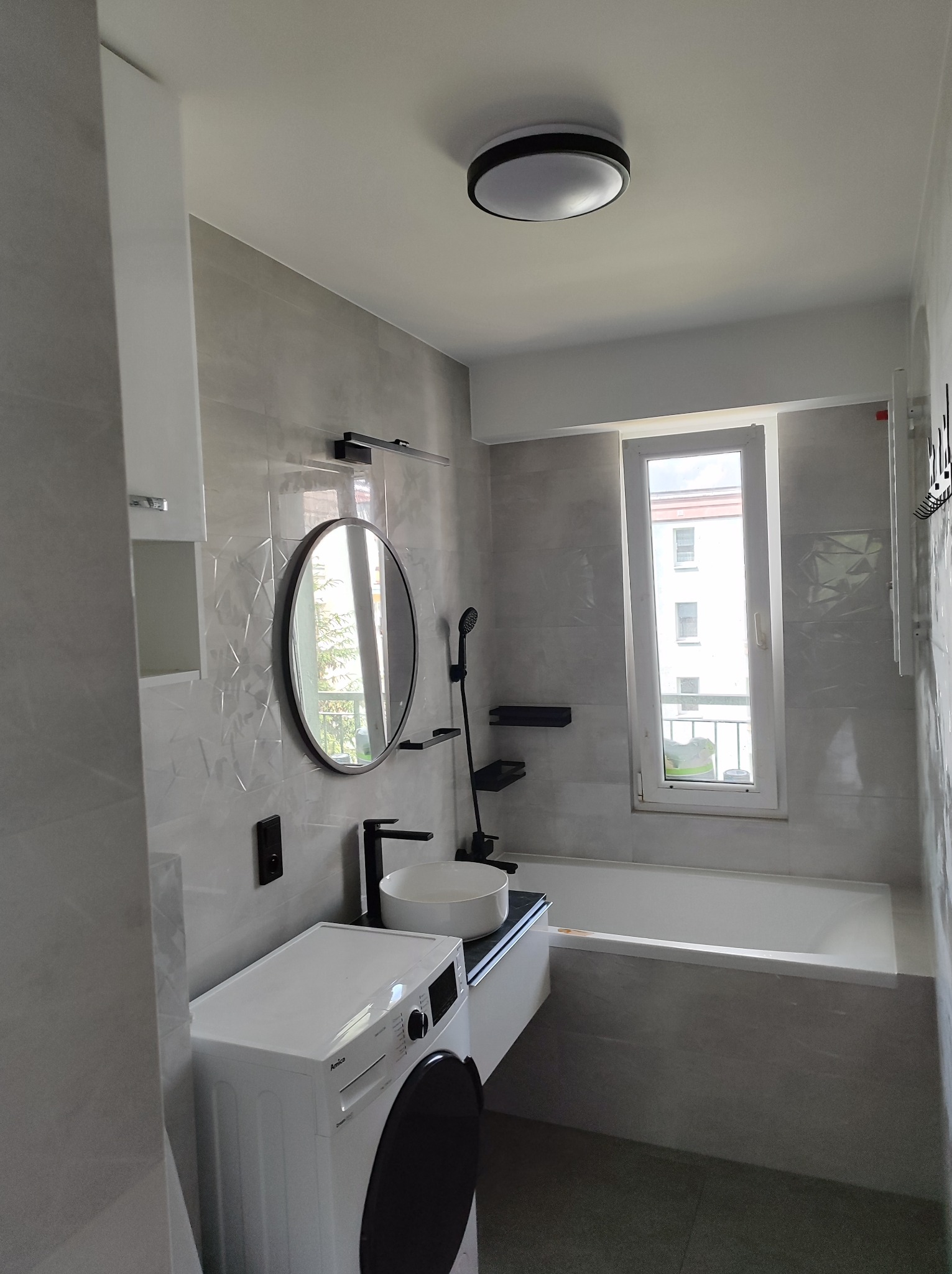 Nowoczesna łazienka z szarymi płytkami, okrągłym lustrem, białą umywalką nablatową, czarną armaturą oraz pralką Amica w rogu.