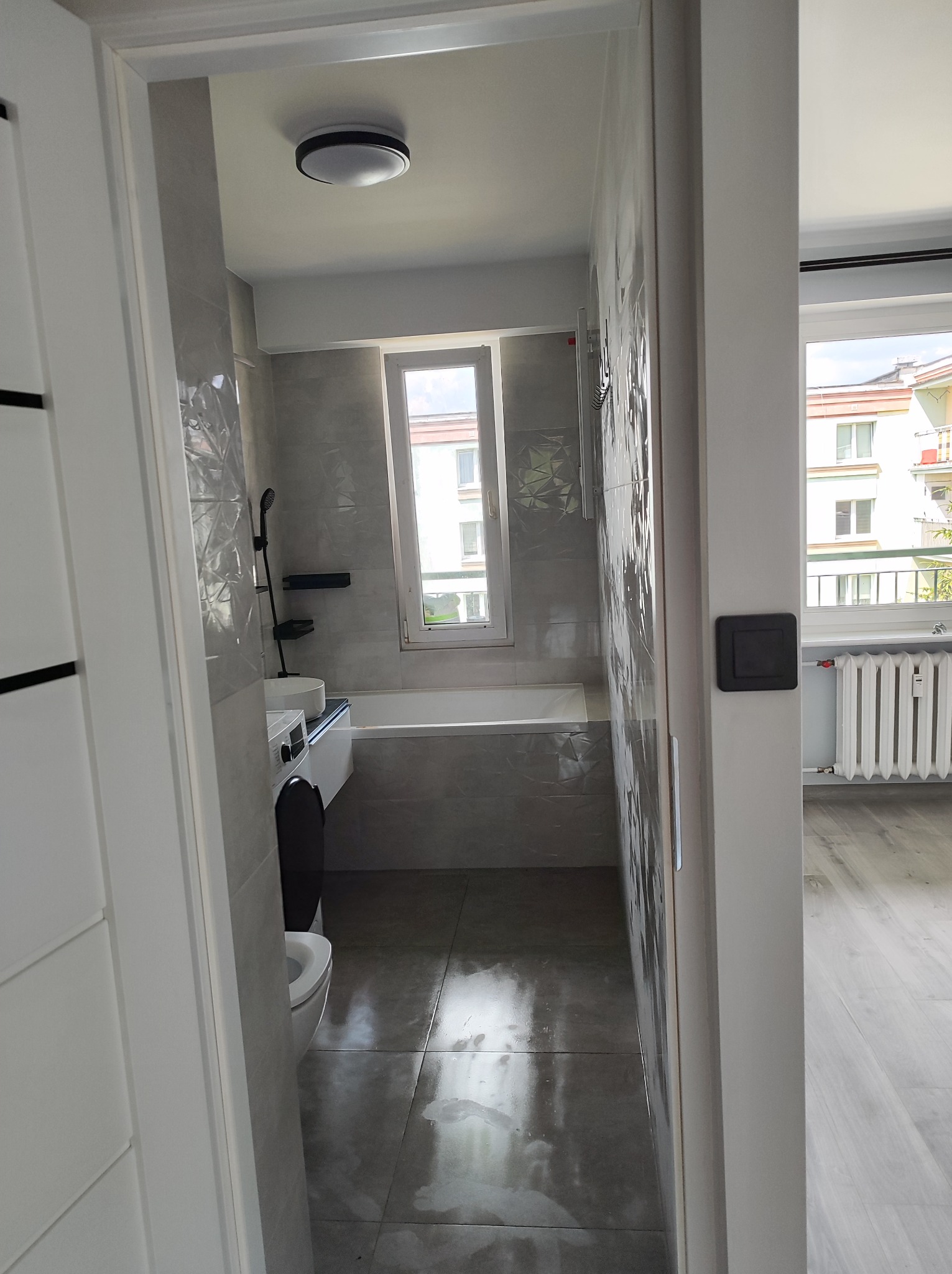 Widok na wyremontowaną łazienkę z szarymi płytkami z wzorem, białą wanną, umywalką i toaletą, widziany przez otwarte drzwi z czarnymi akcentami, z oknem w tle i fragmentem pokoju obok z grzejnikiem.