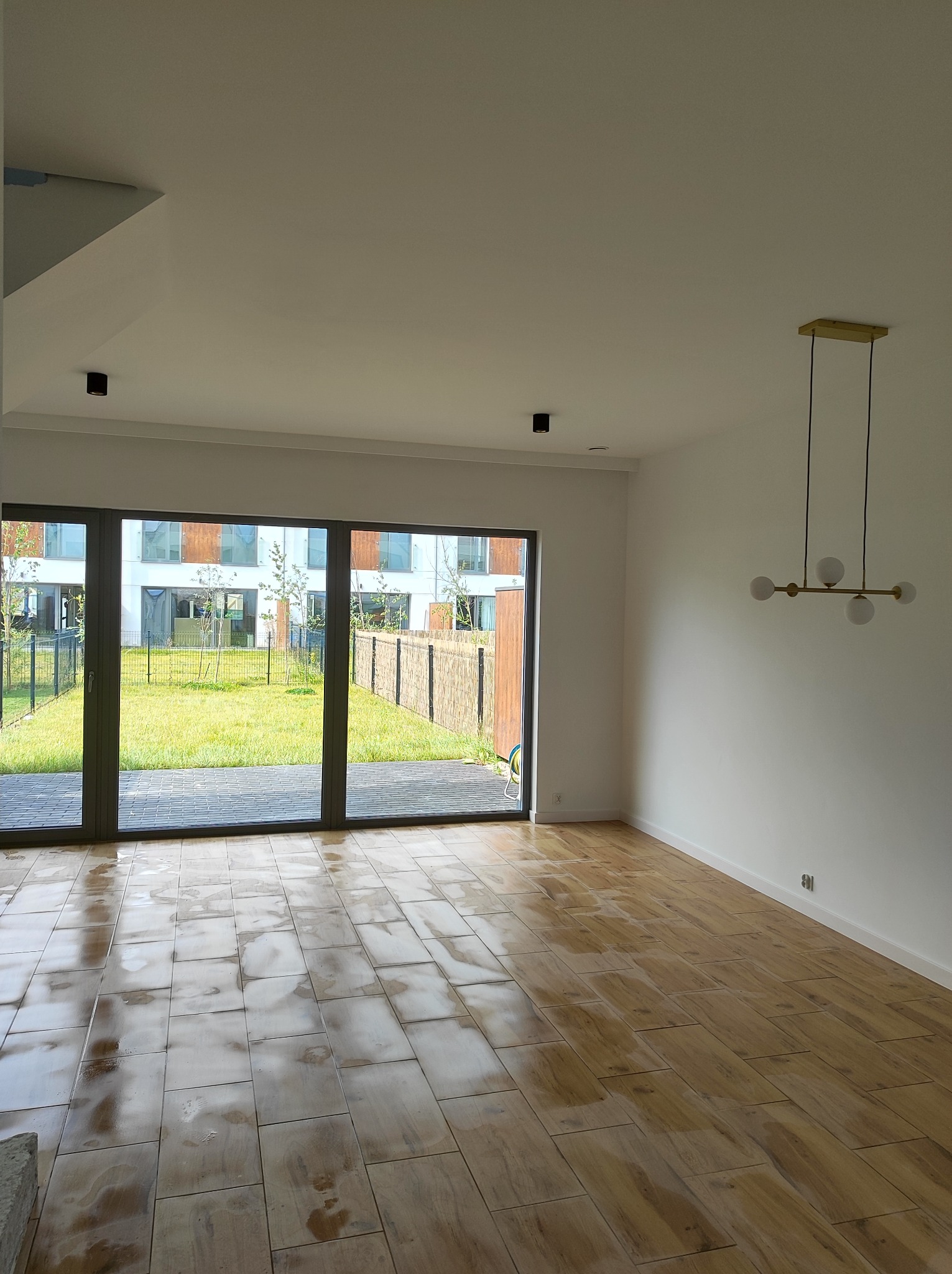 Wnętrze jasnego pokoju z dużym oknem tarasowym wychodzącym na ogród, drewnopodobne płytki podłogowe z widocznymi plamami, nowoczesna lampa wisząca z kulistymi kloszami.