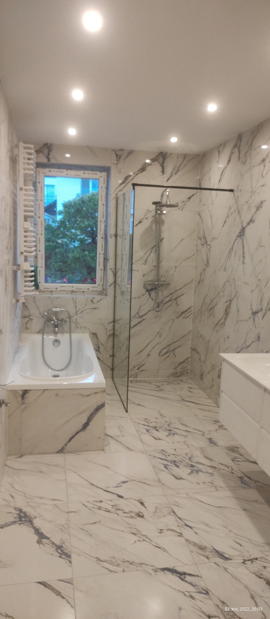 Nowoczesna łazienka z marmurowymi płytkami, przeszkloną kabiną prysznicową i wanną przy oknie, oświetlona punktowymi lampami sufitowymi.