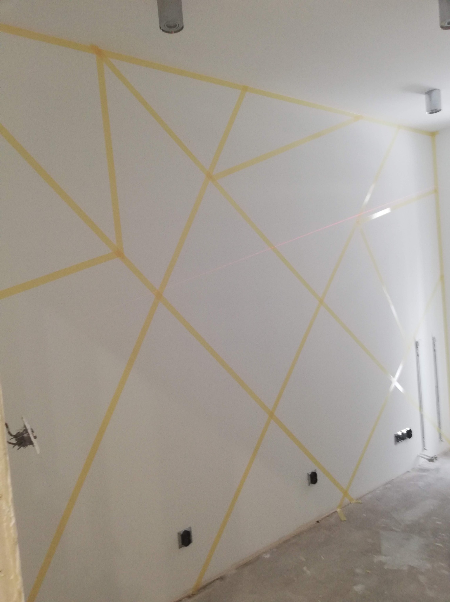 Biała ściana z geometrycznym wzorem wyznaczonym taśmą malarską, widoczne puszki elektryczne i ślady po wiązce lasera, przygotowanie do malowania wzoru.