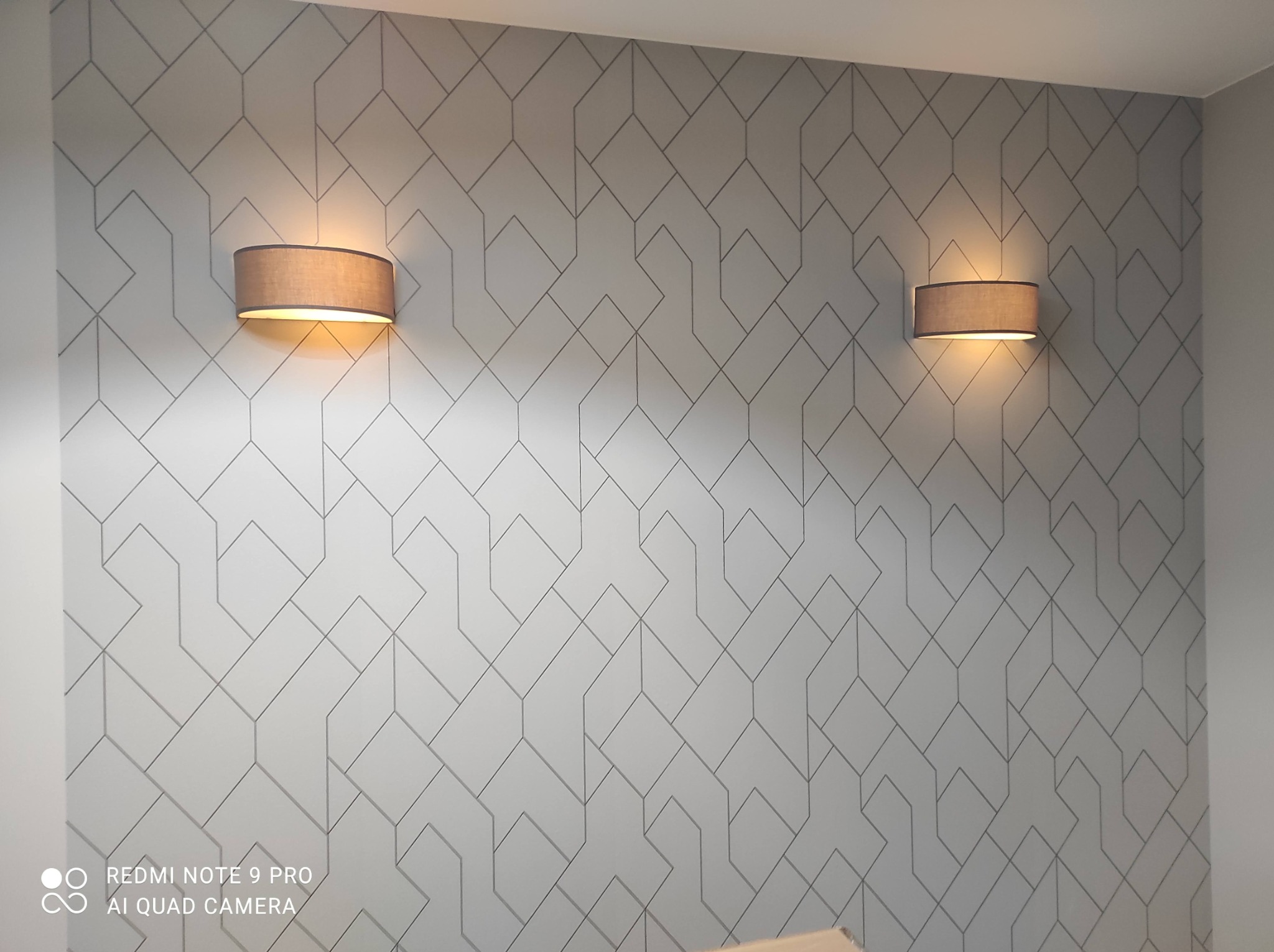Dwa identyczne, brązowe kinkiety z abażurami, emitujące ciepłe światło, zamontowane na ścianie pokrytej geometrycznym, szarym wzorem.