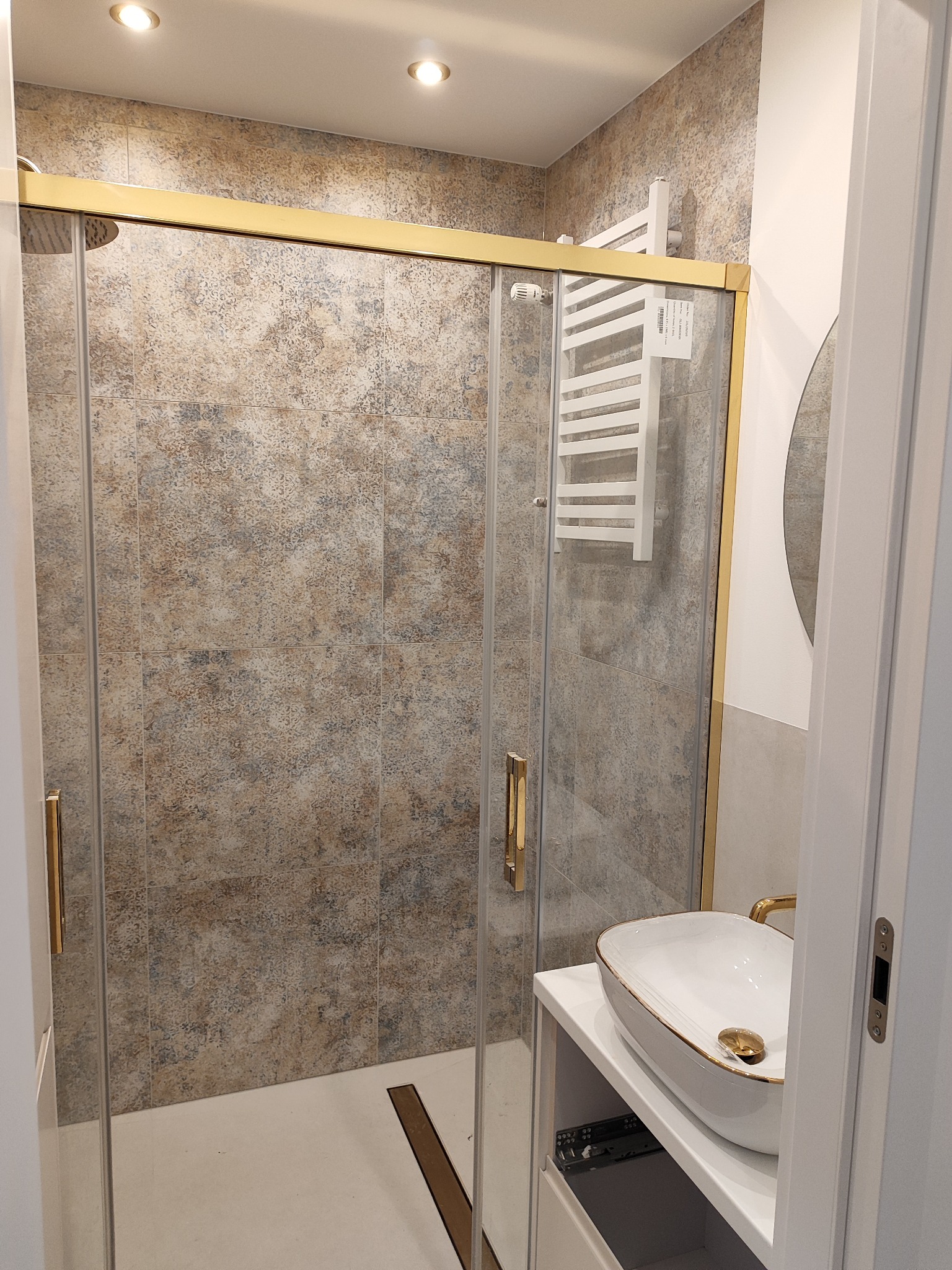 Nowoczesna kabina prysznicowa z przesuwnymi drzwiami w złotej ramie, widoczna umywalka nablatowa z elementami w kolorze złota, biały grzejnik drabinkowy na ścianie wyłożonej płytkami w odcieniach...