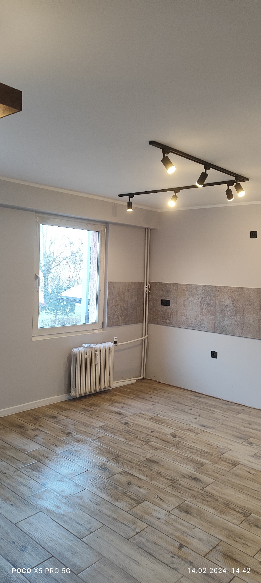 Jasne pomieszczenie po remoncie z panelami podłogowymi imitującymi drewno, grzejnikiem pod oknem i nowoczesnym oświetleniem sufitowym na szynoprzewodach.