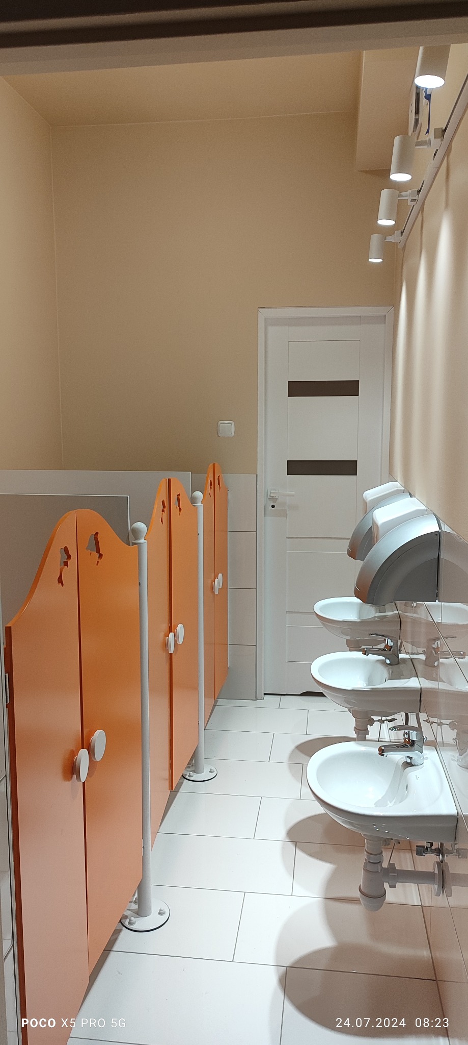 Umywalki dziecięce z suszarkami do rąk w rzędzie, pomarańczowe kabiny toaletowe z wyciętymi motywami zwierzęcymi, białe płytki podłogowe i oświetlenie punktowe na suficie.