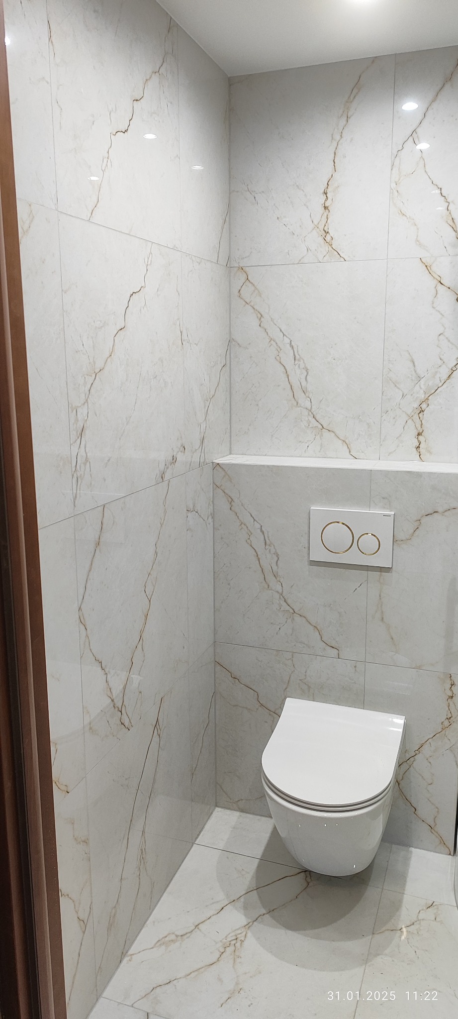 Ściany i podłoga wyłożone białymi płytkami z marmurkowym wzorem w wykończonej łazience, wisząca toaleta z podtynkową spłuczką z dwoma okrągłymi przyciskami w złotym kolorze.