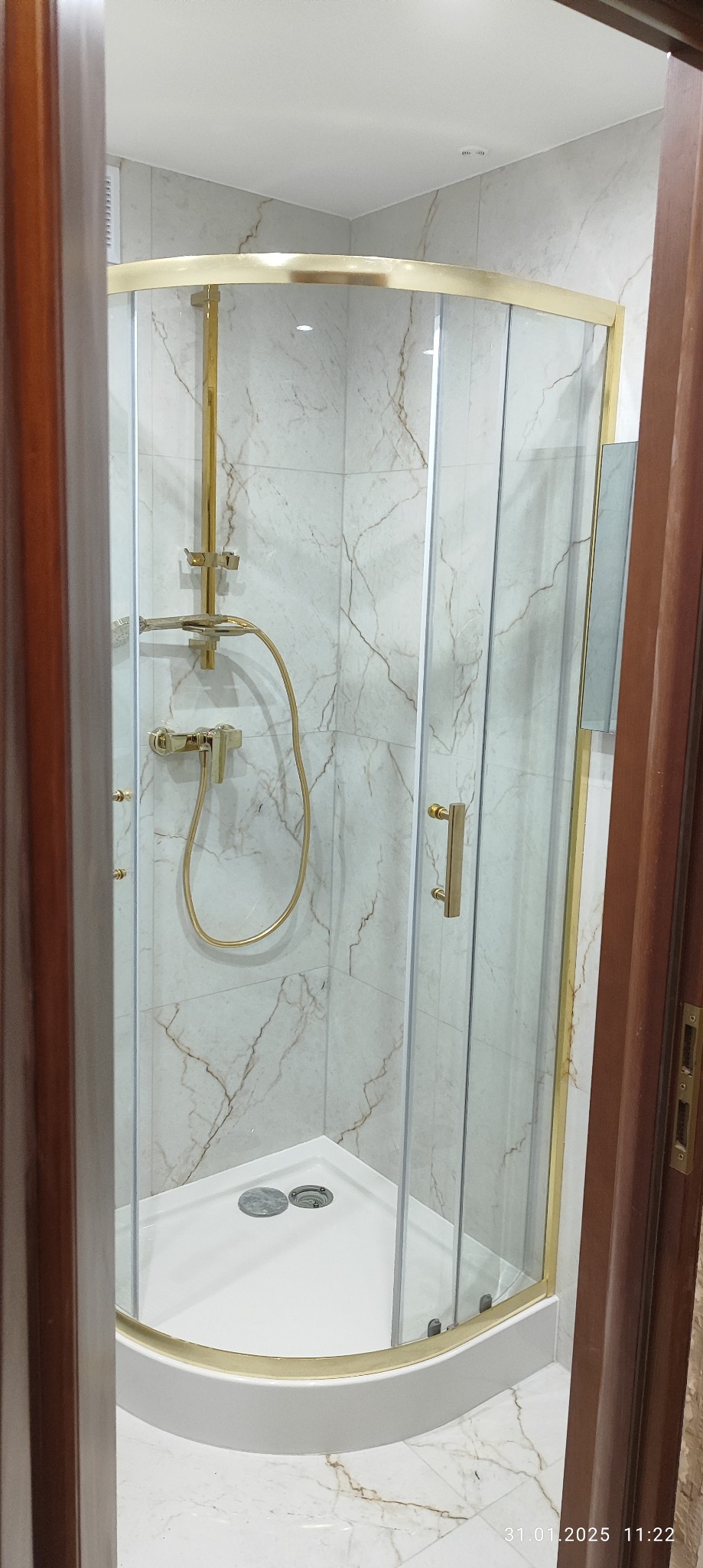 Nowoczesna kabina prysznicowa z marmurowymi płytkami i złotymi detalami, widoczna zza drewnianej framugi drzwi.