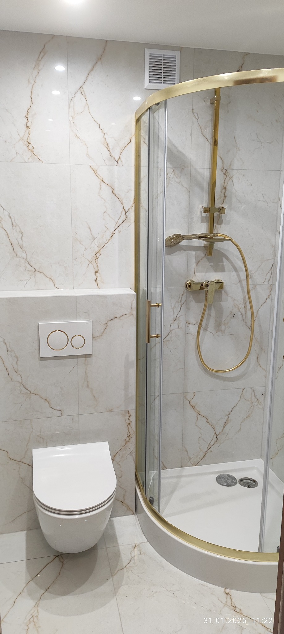 Nowoczesna łazienka z marmurowymi płytkami, kabiną prysznicową z złotymi detalami i podwieszaną toaletą z przyciskiem spłukującym w kolorze białym ze złotymi obwódkami.