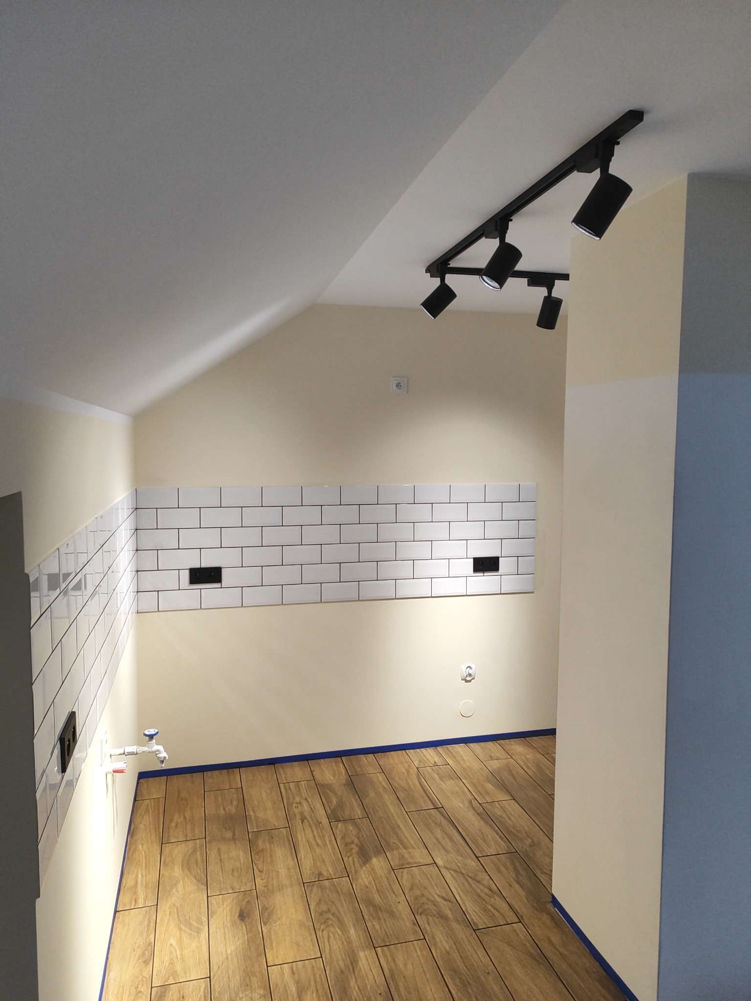 Wykończona łazienka na poddaszu: białe, prostokątne płytki ułożone do połowy ściany, drewnopodobna podłoga z gresu, czarne reflektory sufitowe na szynoprzewodzie.