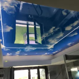 Błękitny sufit z motywem chmur, podświetlony taśmą LED, z odbiciem ogrodu i krzesła w połyskującej powierzchni.