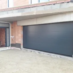 Nowa, antracytowa brama garażowa segmentowa w surowym stanie budynku, z otwartymi drzwiami wejściowymi obok, widoczne niedokończone ściany z cegły i betonowy strop.