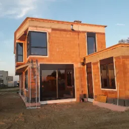 Budynek w trakcie budowy z zamontowanymi oknami z czarnymi ramami, widoczna drabina oparta o ścianę, ziemia wokół budynku.