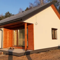 Nowoczesny, parterowy dom z białymi ścianami i drewnianą elewacją, z ciemnymi oknami i dachem, widoczny na tle lasu, z paletami na tarasie i zwisającym kablem na elewacji.