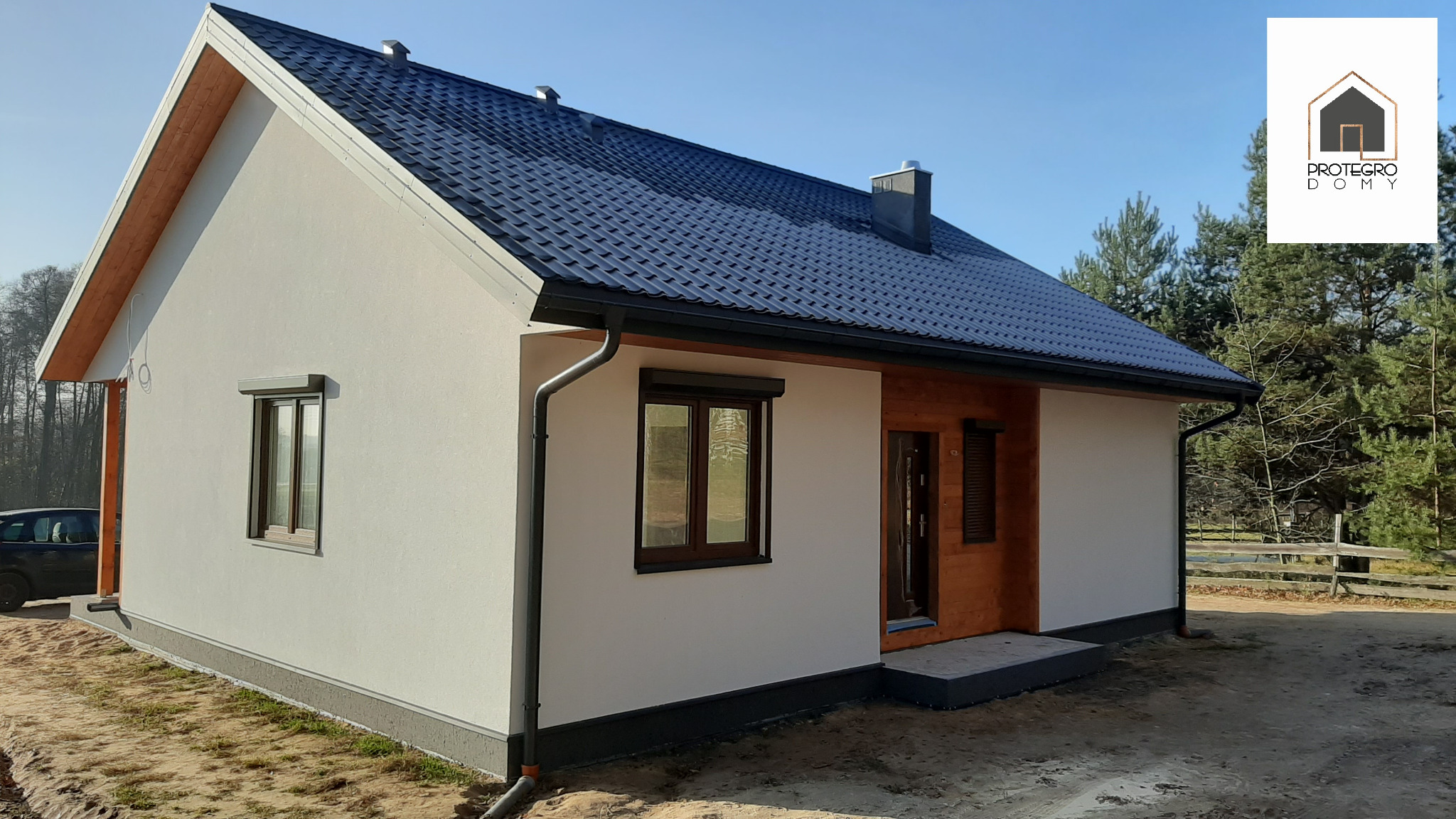 Gotowy dom jednorodzinny z ciemną dachówką i jasną elewacją, widoczne okna z roletami zewnętrznymi, drewniane elementy wykończeniowe i logo firmy w prawym górnym rogu.