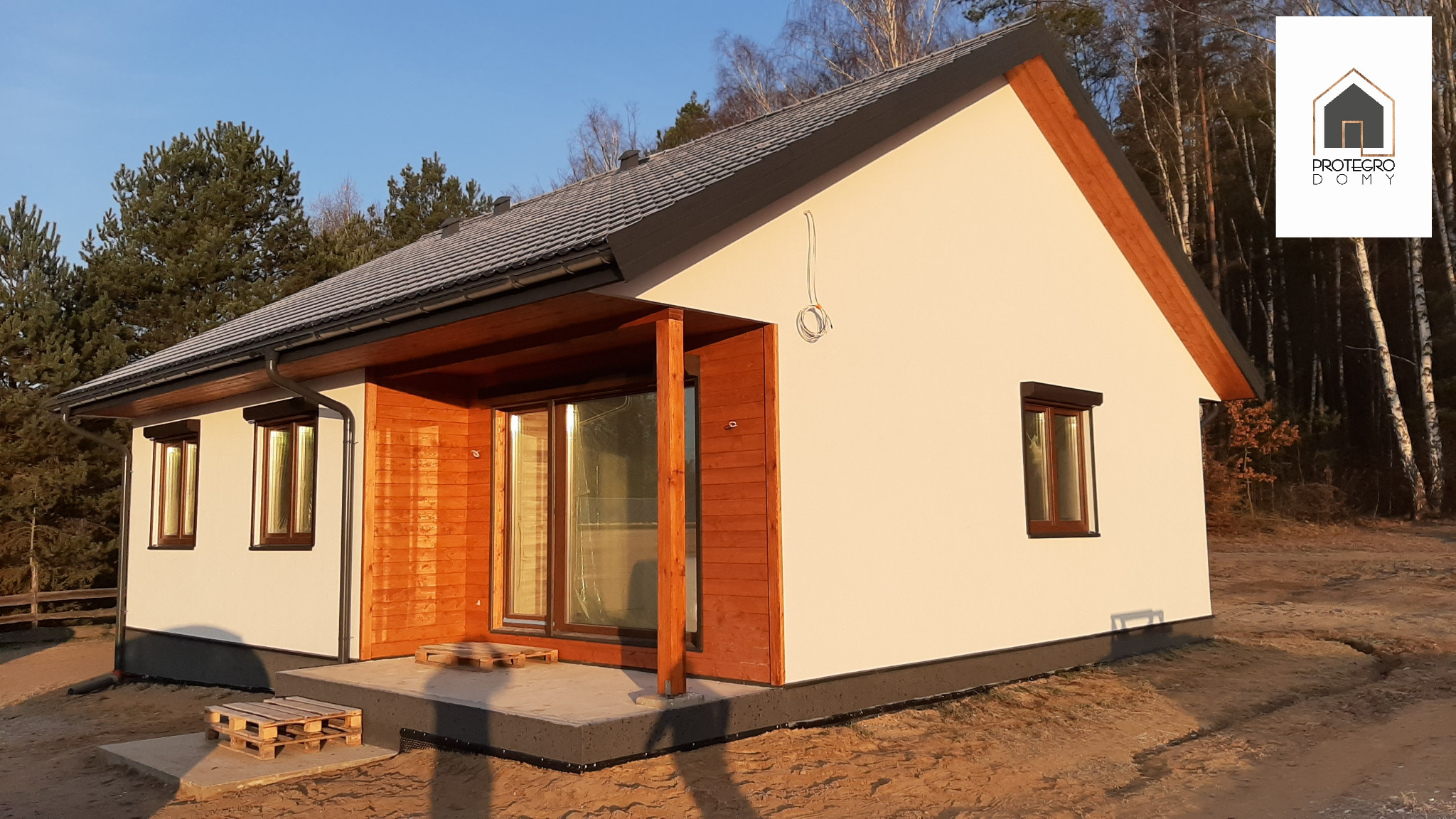 Nowoczesny, parterowy dom z białymi ścianami i drewnianą elewacją, z ciemnymi oknami i dachem, widoczny na tle lasu, z paletami na tarasie i zwisającym kablem na elewacji.