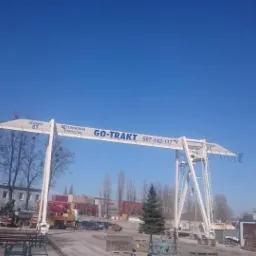 Biały suwnicowy dźwig bramowy z napisem 'GO-TRAKT' i numerem telefonu na tle błękitnego nieba, drzew bez liści i budynku z szarymi ścianami, z widocznymi blokami betonowymi i elementami...