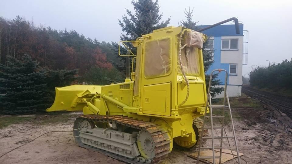 Żółty spychacz gąsienicowy po malowaniu, z kabiną zabezpieczoną folią, stojący na placu budowy z widoczną drabiną i budynkiem w tle.