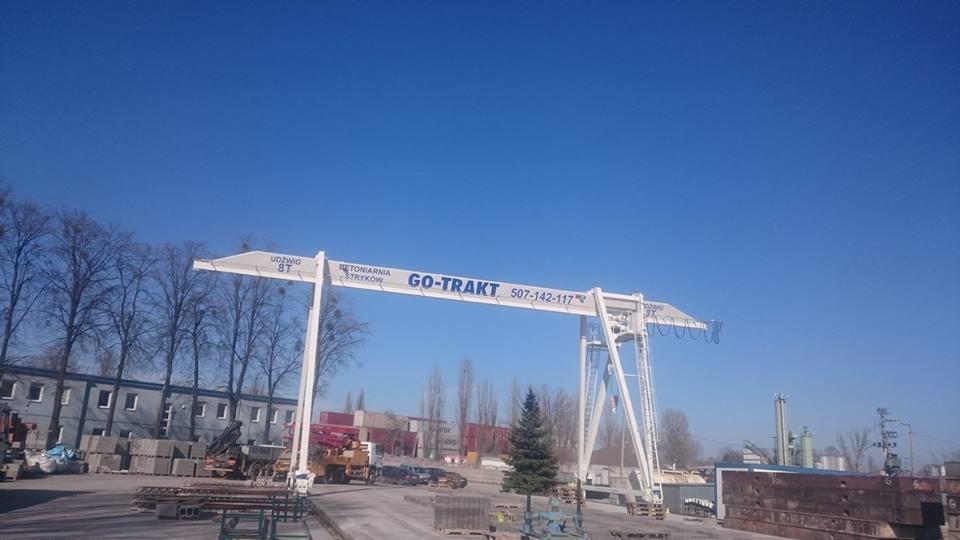 Biały suwnicowy dźwig bramowy z napisem 'GO-TRAKT' i numerem telefonu na tle błękitnego nieba, drzew bez liści i budynku z szarymi ścianami, z widocznymi blokami betonowymi i elementami...