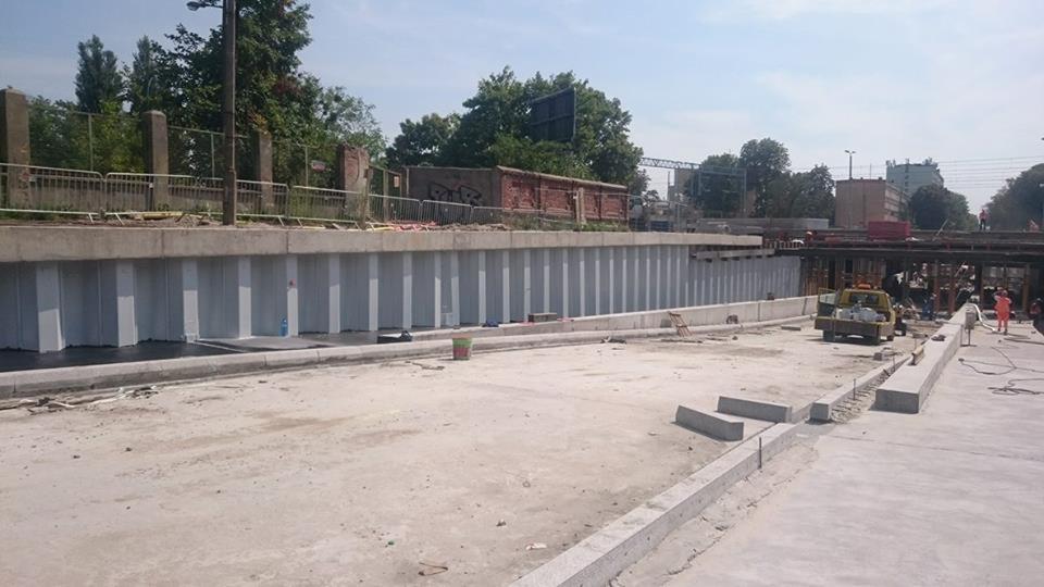 Konstrukcja betonowa z pionowymi panelami, prawdopodobnie ściana oporowa, w trakcie budowy; widoczne elementy szalunku, betonowe krawężniki, sprzęt budowlany i pracownicy w oddali.