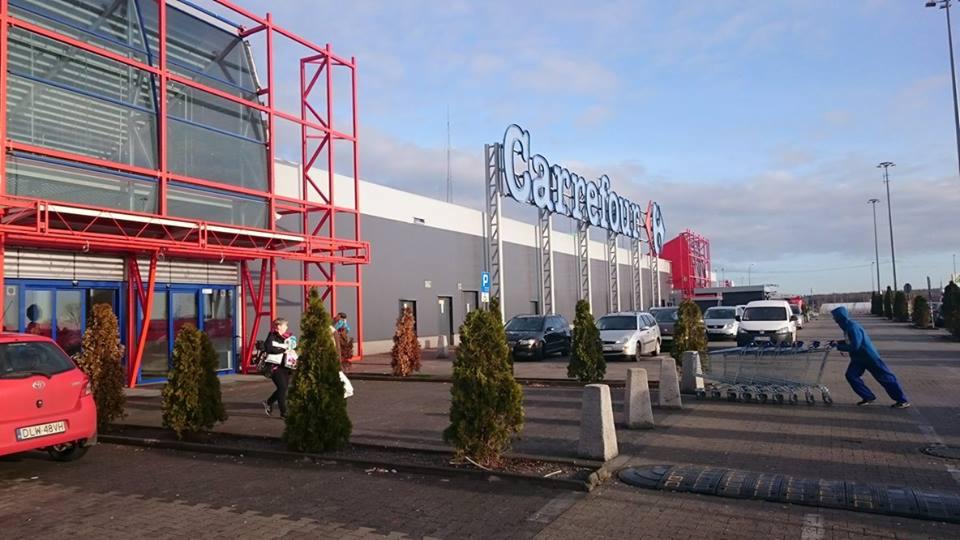 Zewnętrzny widok na budynek sklepu Carrefour z czerwonymi elementami konstrukcyjnymi, parkingiem, zielenią i osobą w niebieskim kombinezonie pchającą wózki sklepowe.