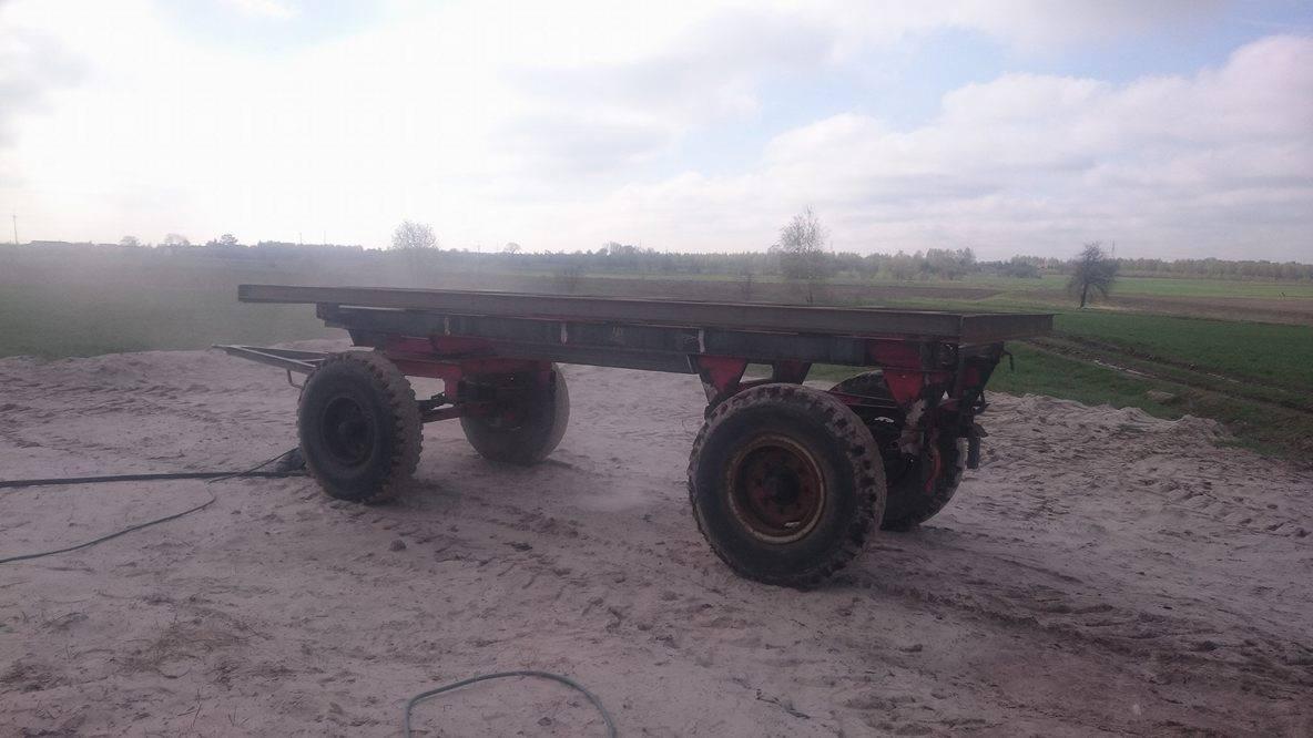 Przyczepa rolnicza z metalową konstrukcją na piaszczystym podłożu, widok na pole i pochmurne niebo w tle.